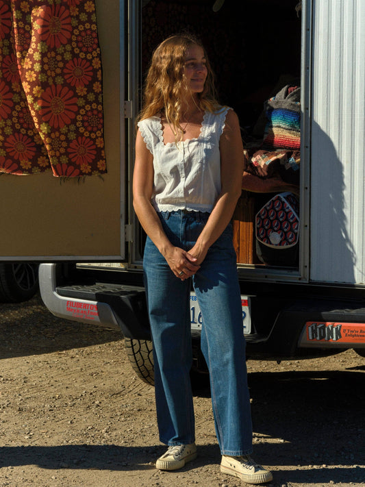 Woman in a light cotton top and denim jeans stands by an open van with sunlit blankets and woven decor inside.