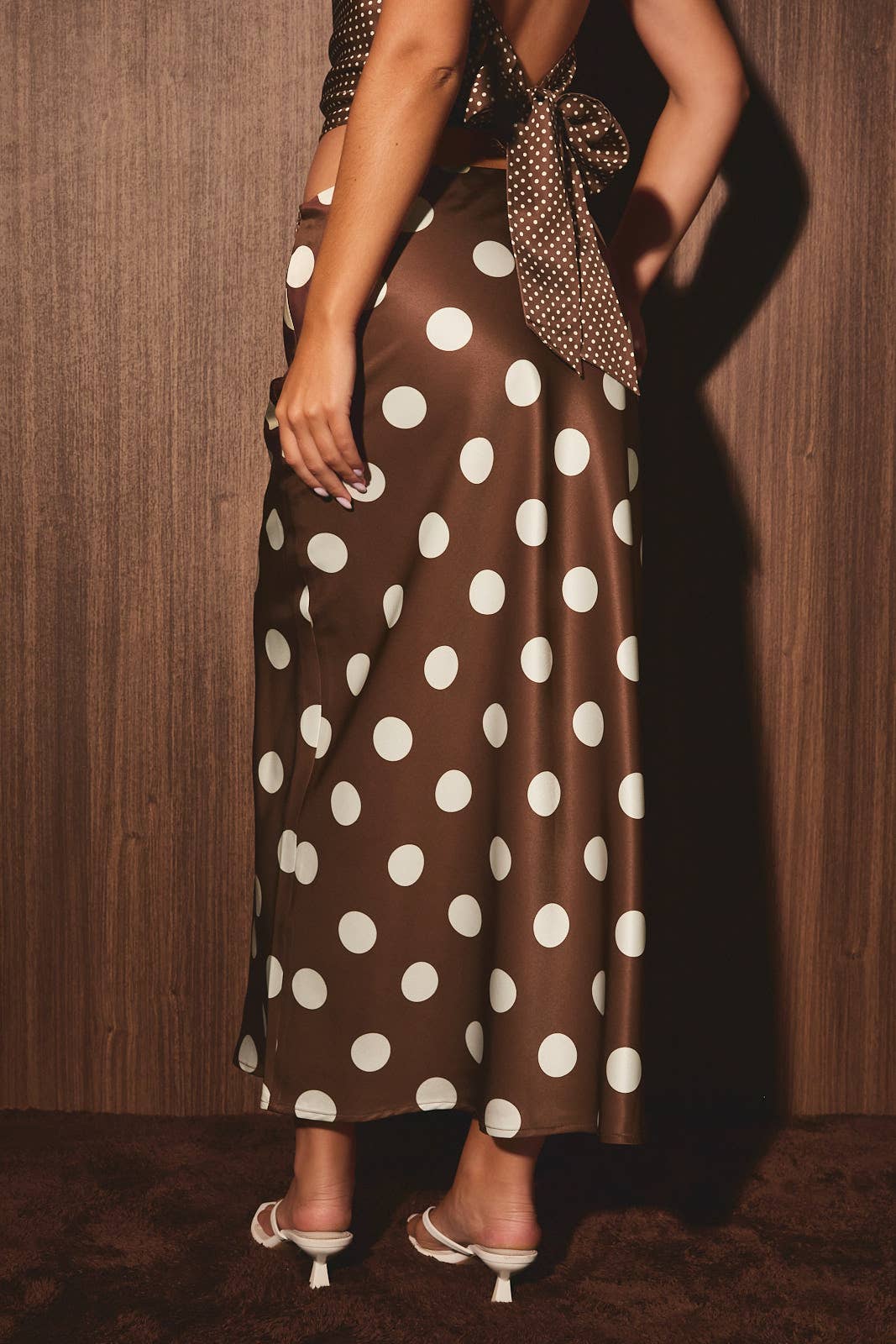 Woman wearing a matching skirt set and white heels poses near a wooden wall, turned slightly to the side.