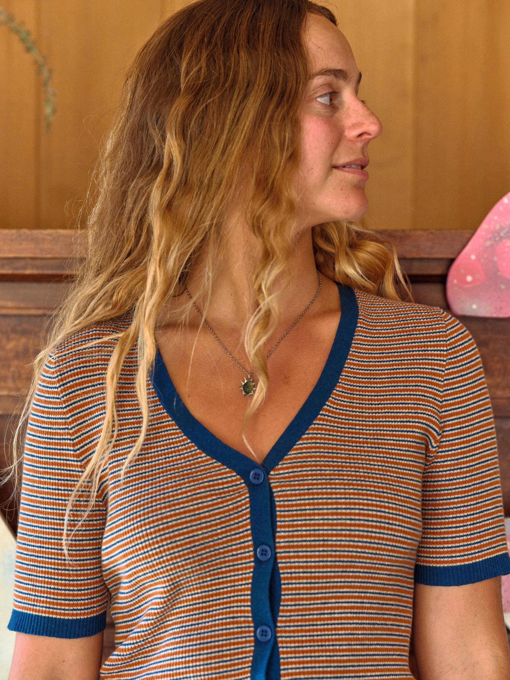 Woman with wavy hair wears a striped cotton tee indoors, standing next to a wooden wall.