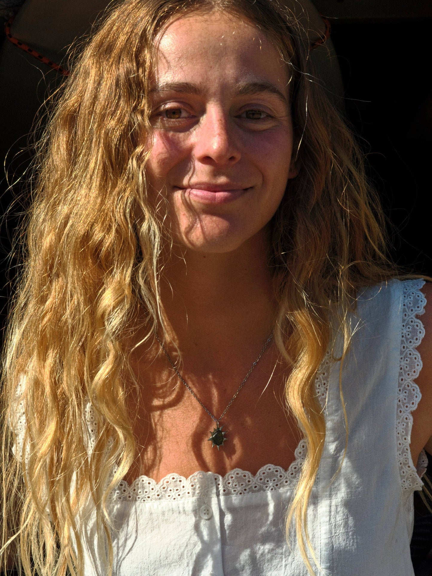 Woman wearing sleeveless linen Helena top and star necklace, standing in sunlight with wavy hair.