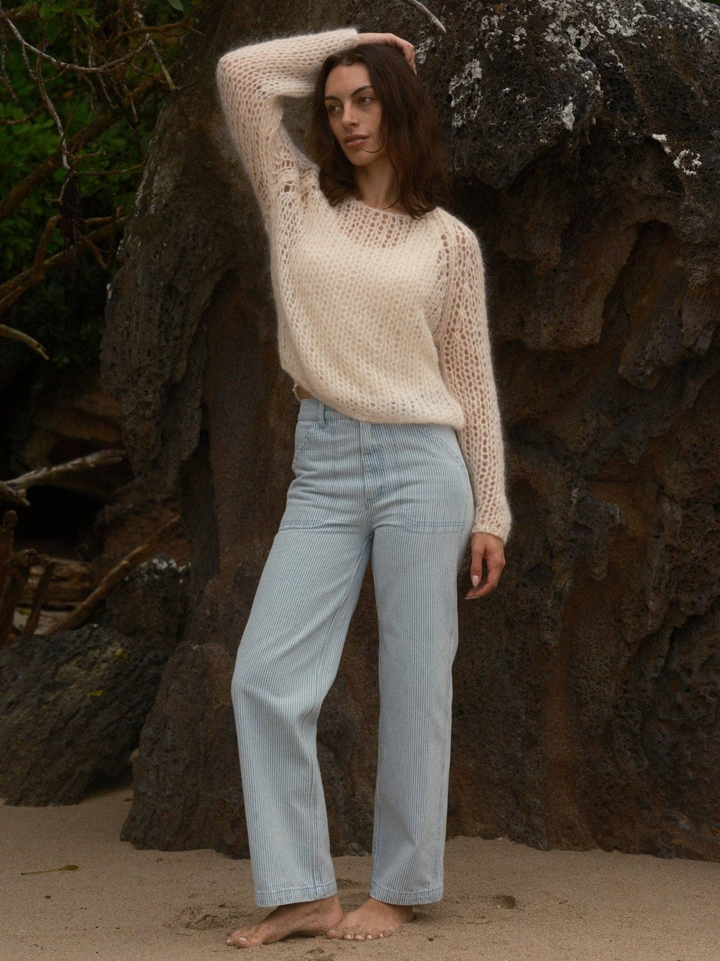 Woman in a cream knit sweater and blue jeans, barefoot on sand, standing before large, rugged rocks.