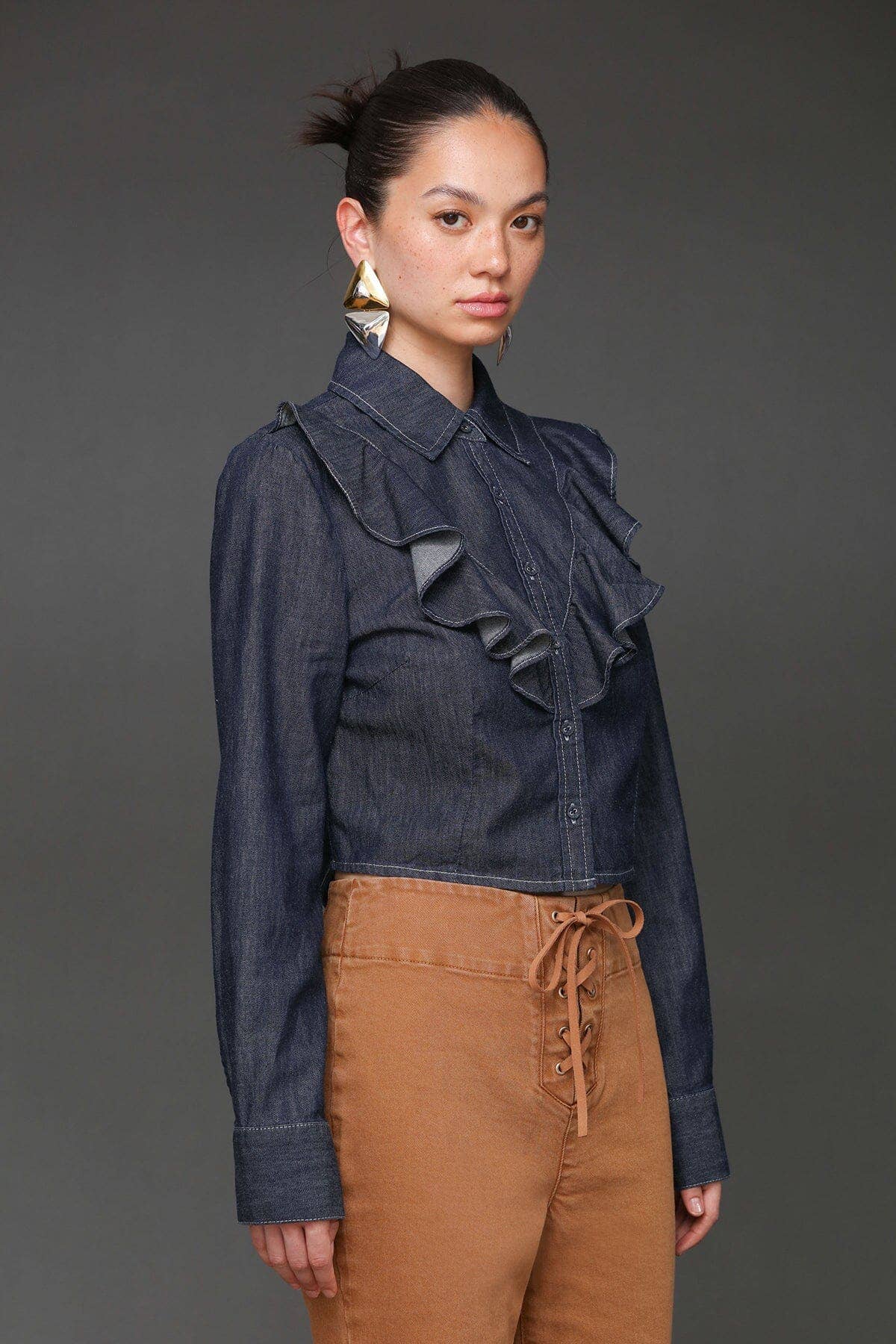 Woman wearing denim cropped shirt and high-waisted tan lace-up pants, standing against a dark backdrop.