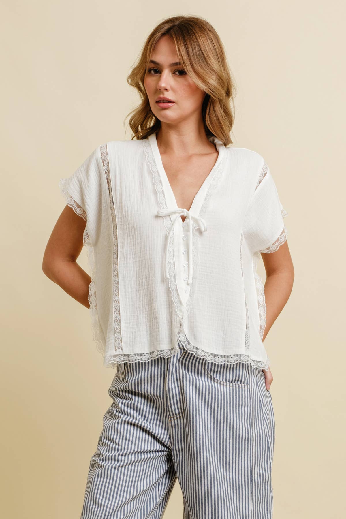 Woman in a lace-trimmed white top and striped pants, standing against a neutral beige background.