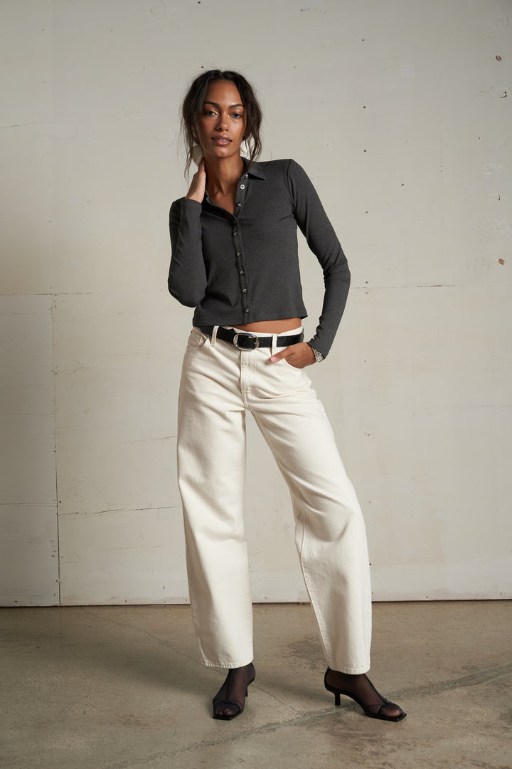 Woman wears a white button-up tee, white wide-leg jeans, and heels, standing indoors against a plain wall.
