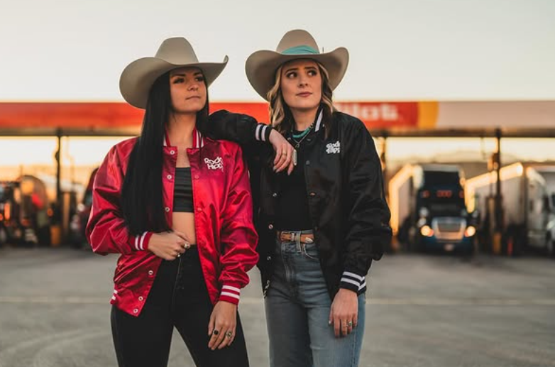 Two women in satin country club jackets stand by trucks at a gas station, both looking to the side.