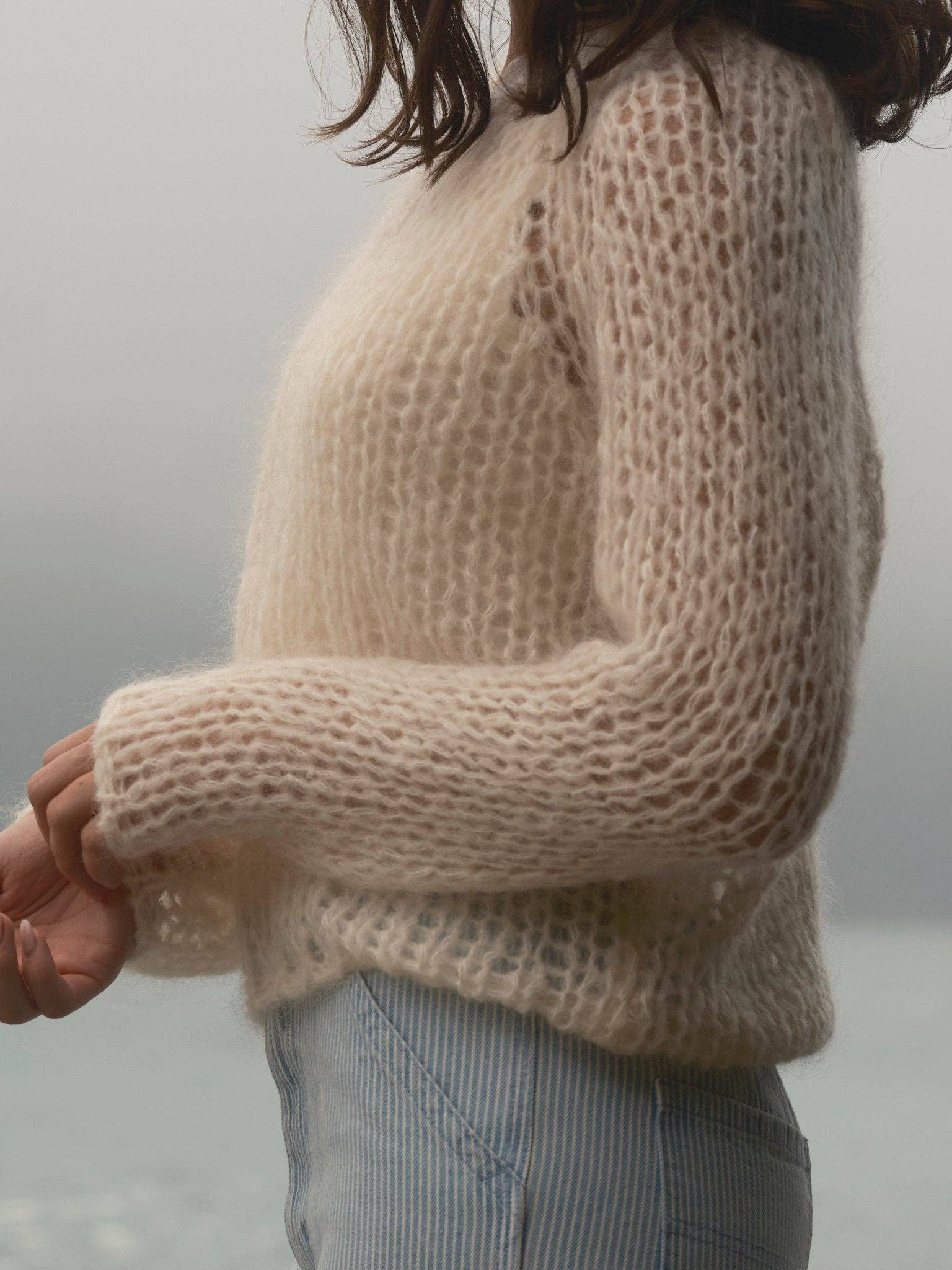 Cream knit sweater paired with striped pants; woman stands by water under a cloudy sky. Sweater is cozy and casual.