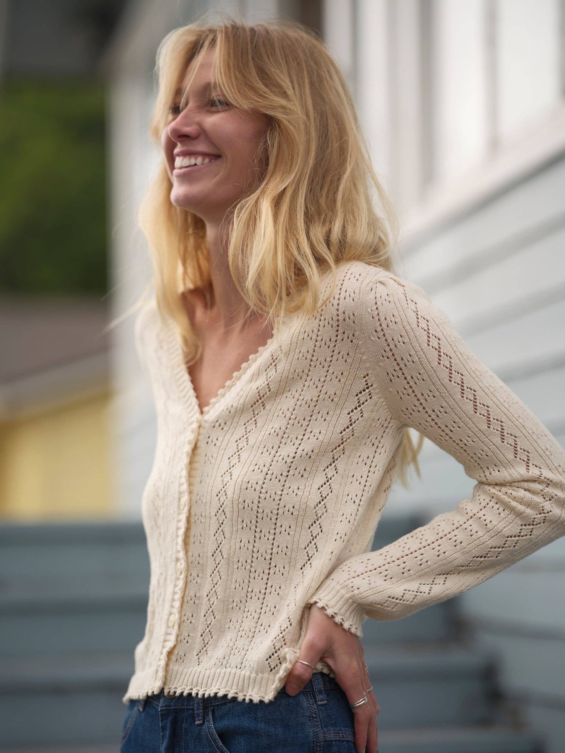Woman with long blonde hair wears a cream knit cardigan, standing outdoors by blue stairs.