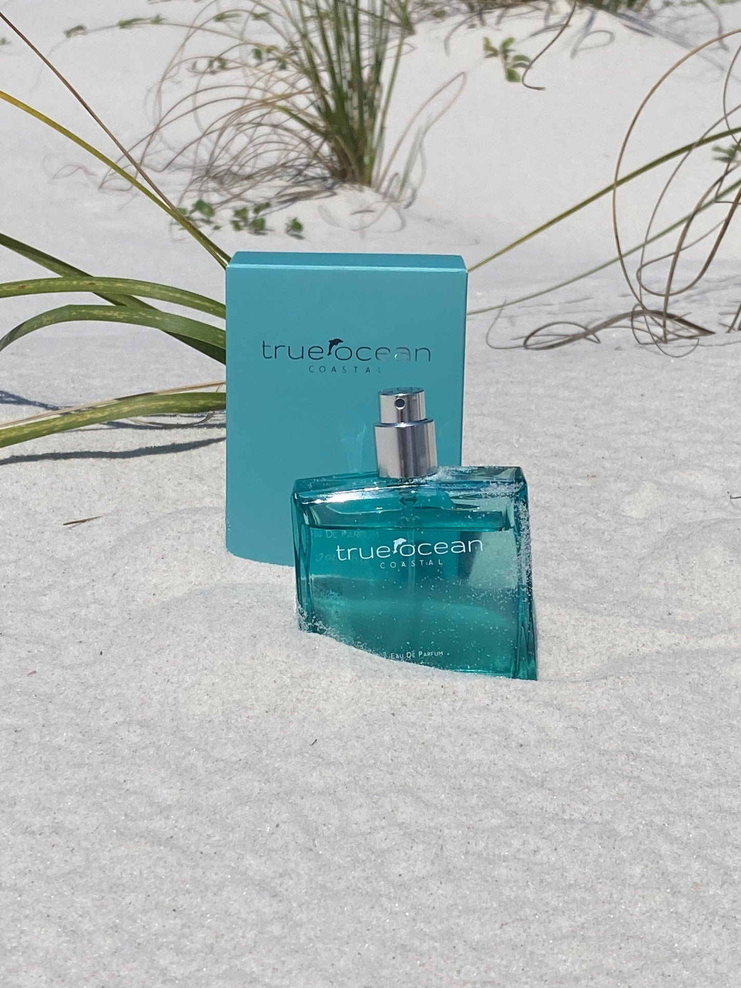 Clear glass perfume bottle and box on white sand, with green beach grass behind. Suitable for personal fragrance use.