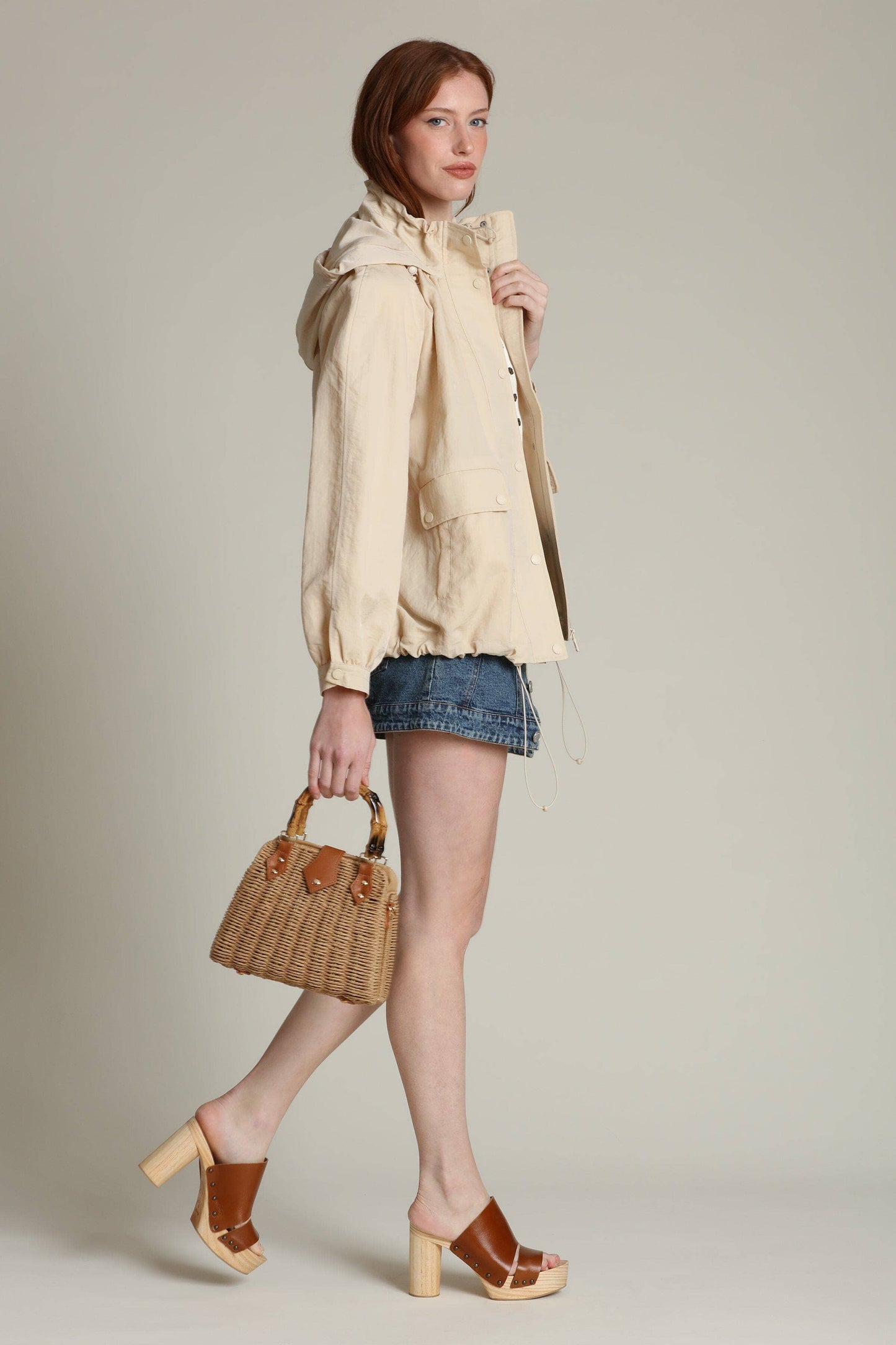 Woman wears a balloon-sleeve windbreaker, denim shorts, and heels, holding a woven bag against a plain background.