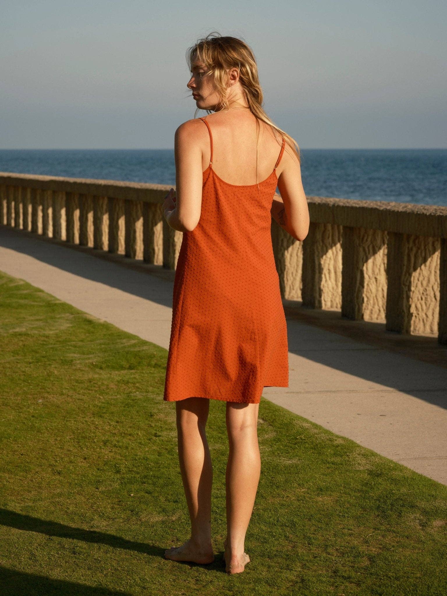 Barefoot woman in a light-colored dobby slip dress stands on grass beside stone railing, overlooking the sea.