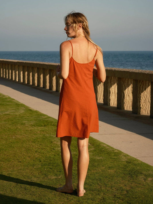 Barefoot woman in a light-colored dobby slip dress stands on grass beside stone railing, overlooking the sea.
