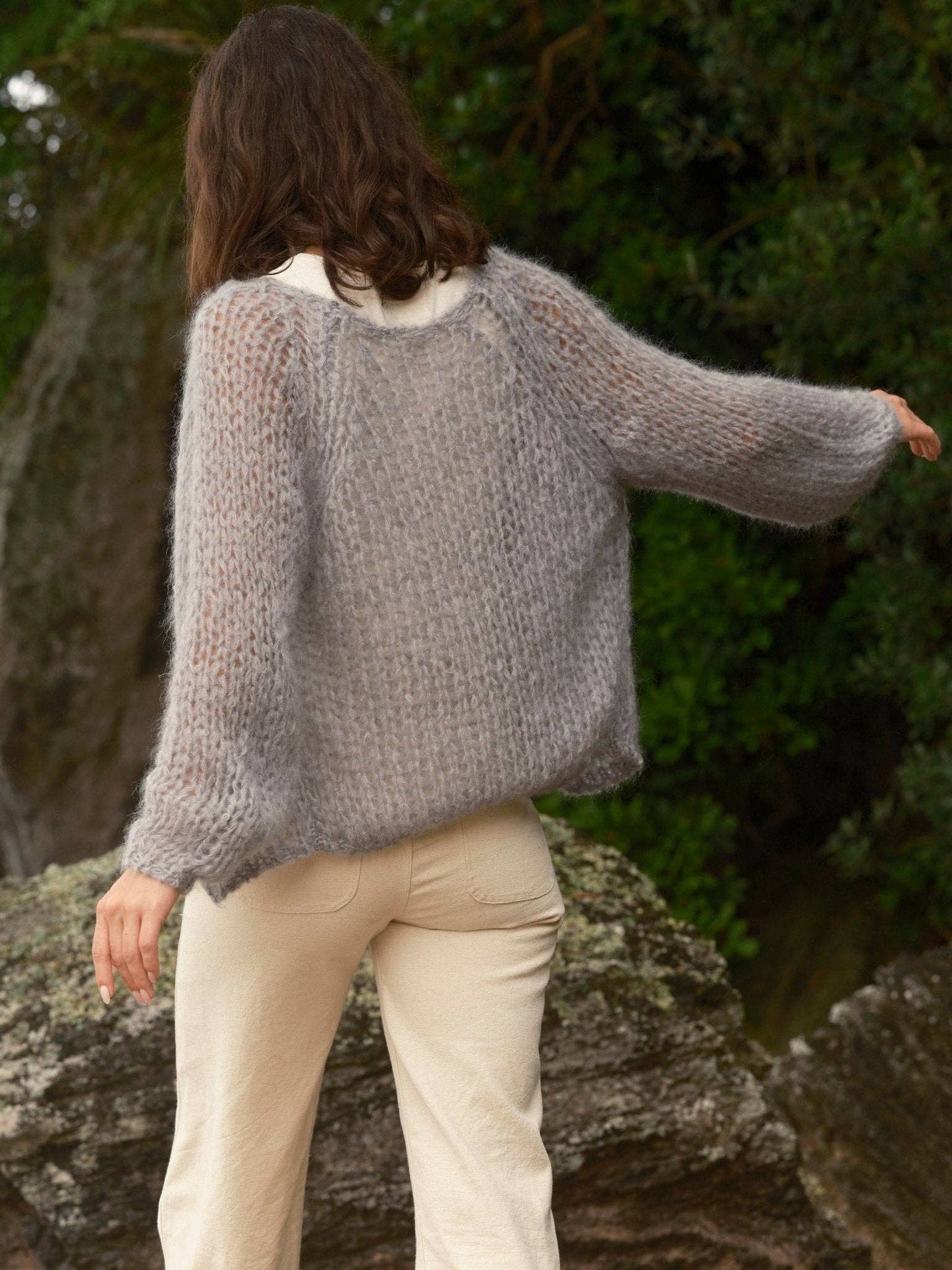 Individual outside wearing a gray wool cardigan and cream pants, shown from behind with one arm outstretched.