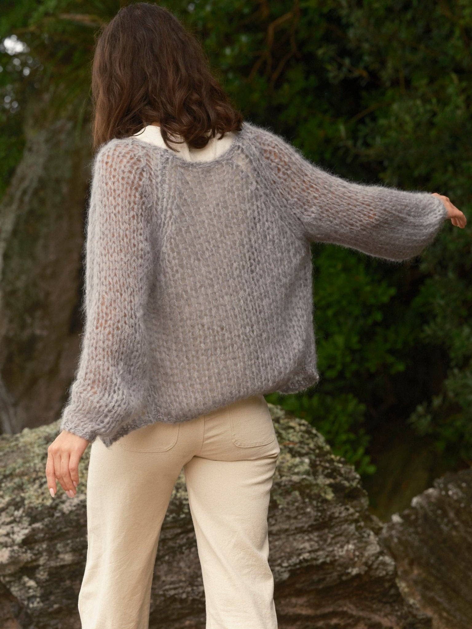 Individual outside wearing a gray wool cardigan and cream pants, shown from behind with one arm outstretched.