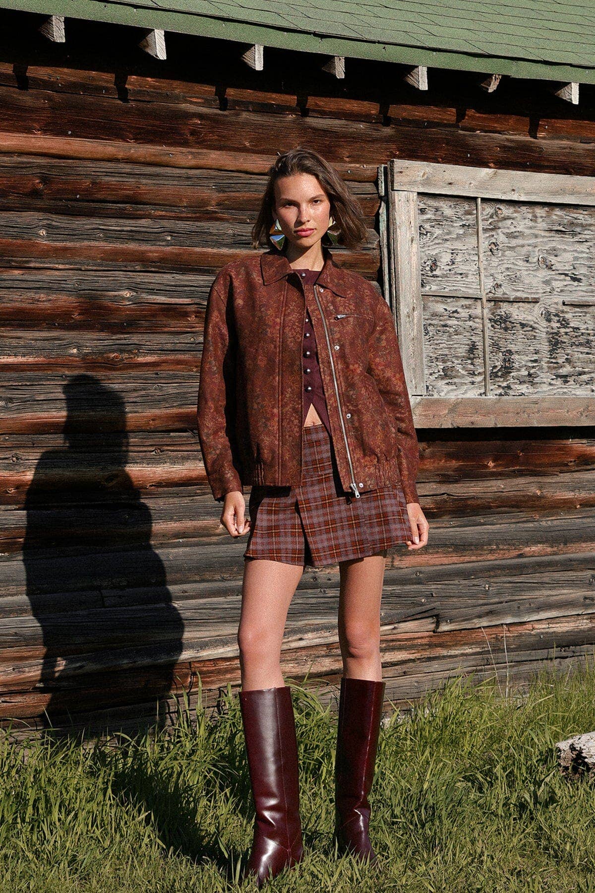 Woman wears a floral print bomber jacket in lightweight fabric, standing outside in front of a weathered wooden building.