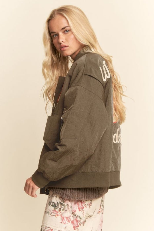 Woman with long blonde hair models an embroidered bomber jacket, featuring western motifs on dark fabric, shown from behind.