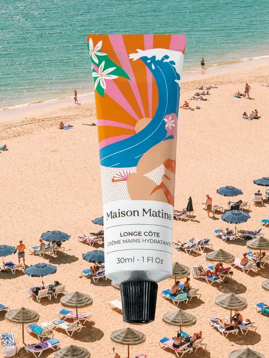 White Maison Matine Longe Côte 30ml hand cream tube hovers above a crowded, sunny beach with umbrellas and people.