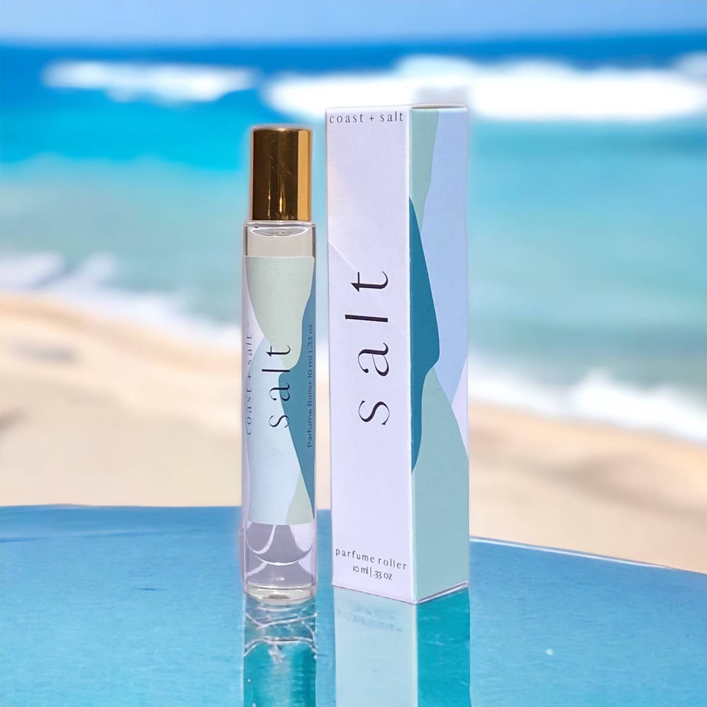 Clear glass perfume roller and matching box on a reflective surface, suggesting scents of sea salt and driftwood.