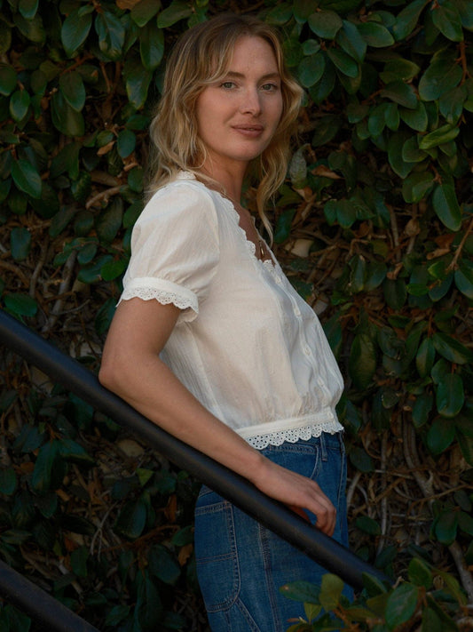 Woman wears a light Lucia top and blue jeans, standing by a staircase with green plants in the background.