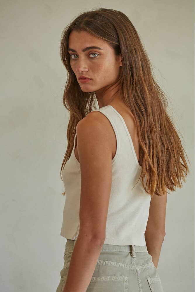 Woman with long brown hair models a white ribbed tank top and light pants, shown from behind against a plain background.