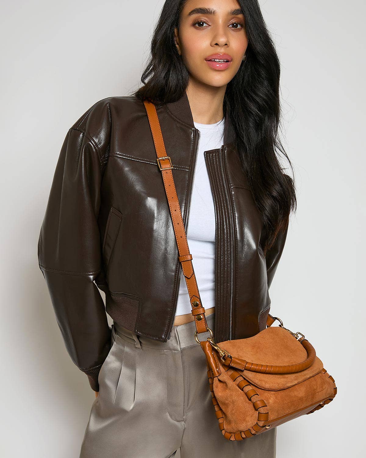 Woman in brown leather jacket, tan pants, carrying a tan leather convertible crossbody bag against a white backdrop.