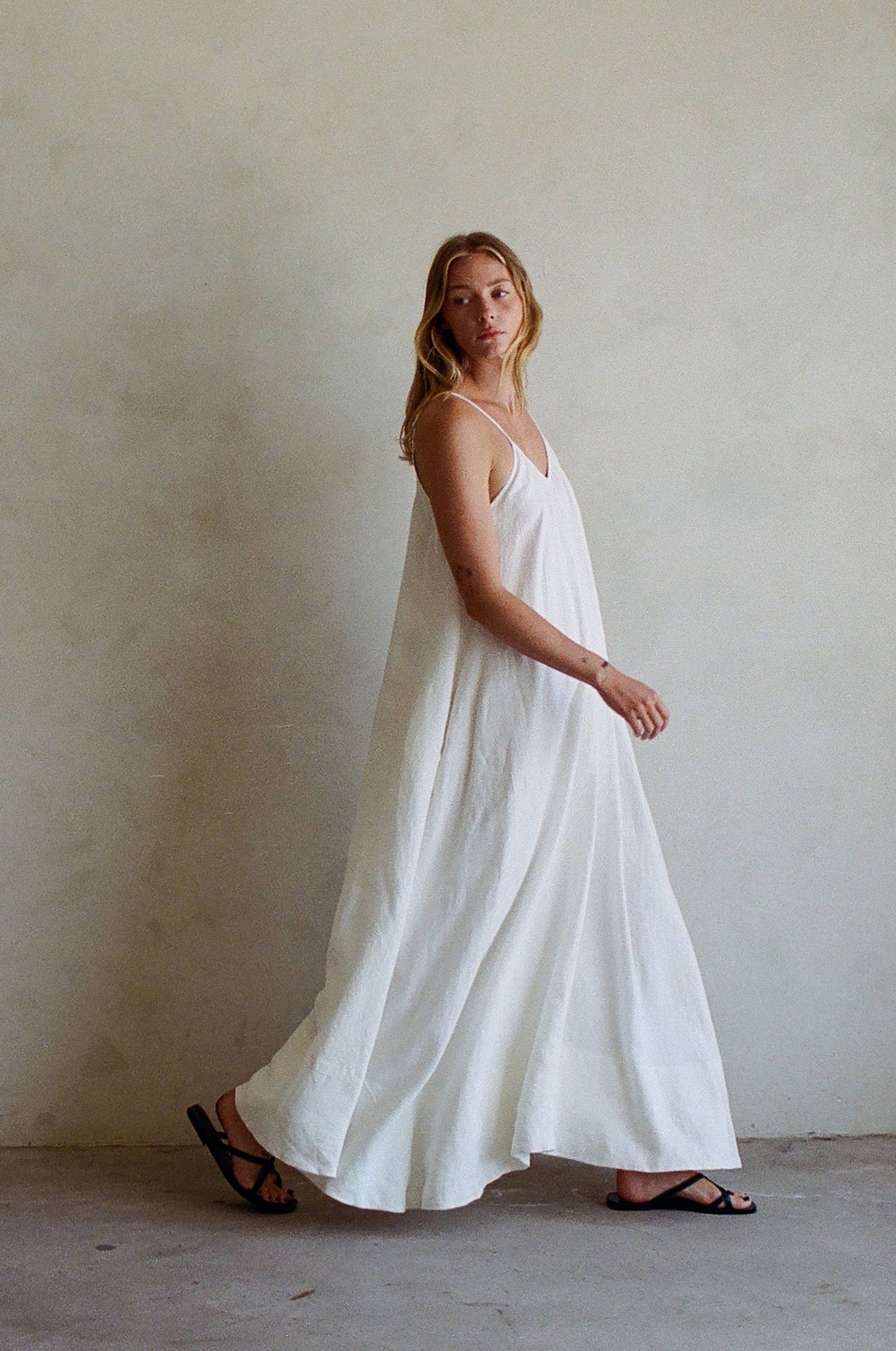 Woman in a linen maxi dress with black sandals walking in front of a plain, light background.