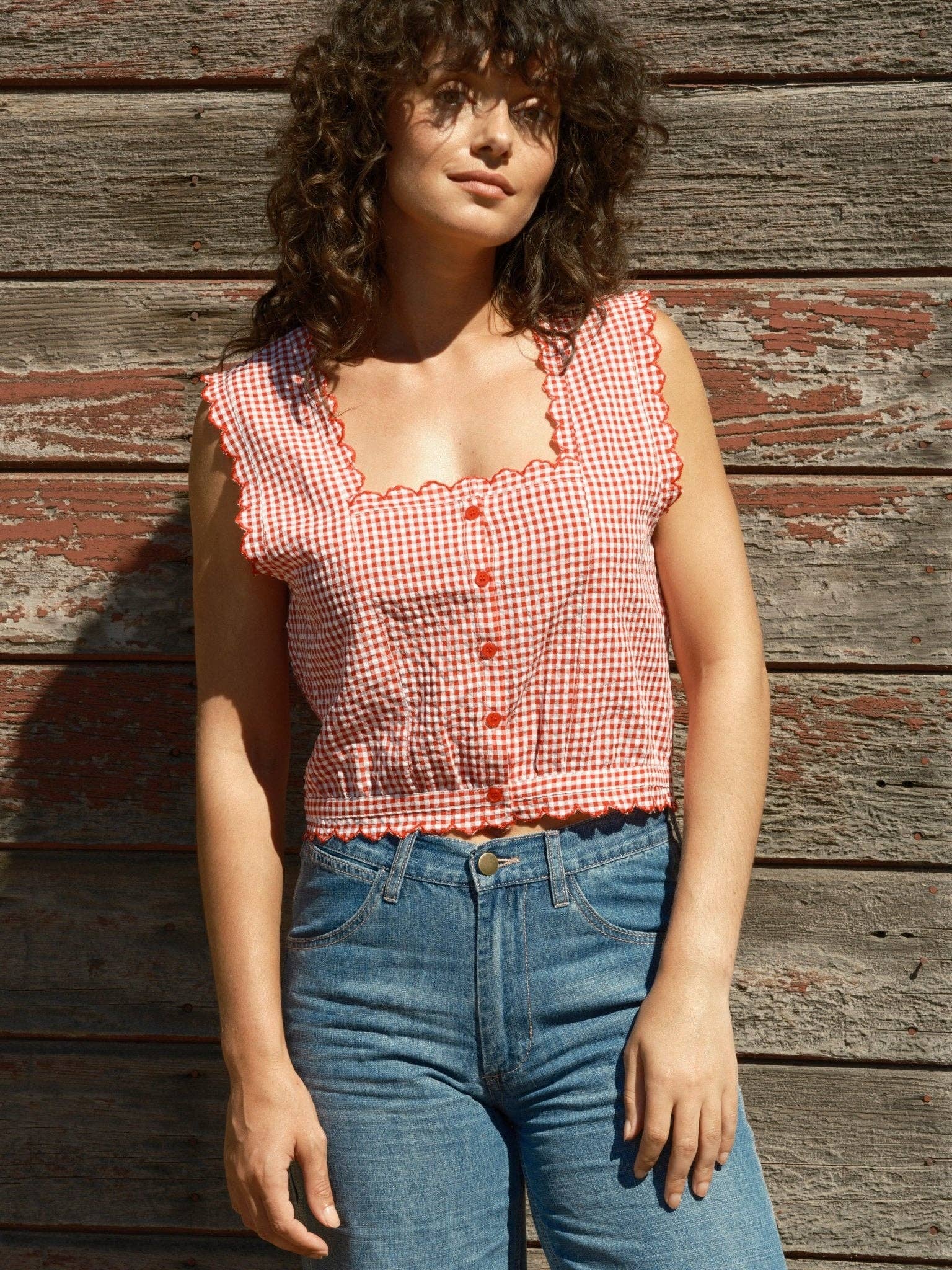 Sleeveless gingham square-neck top with blue jeans, worn by a rustic wooden wall.