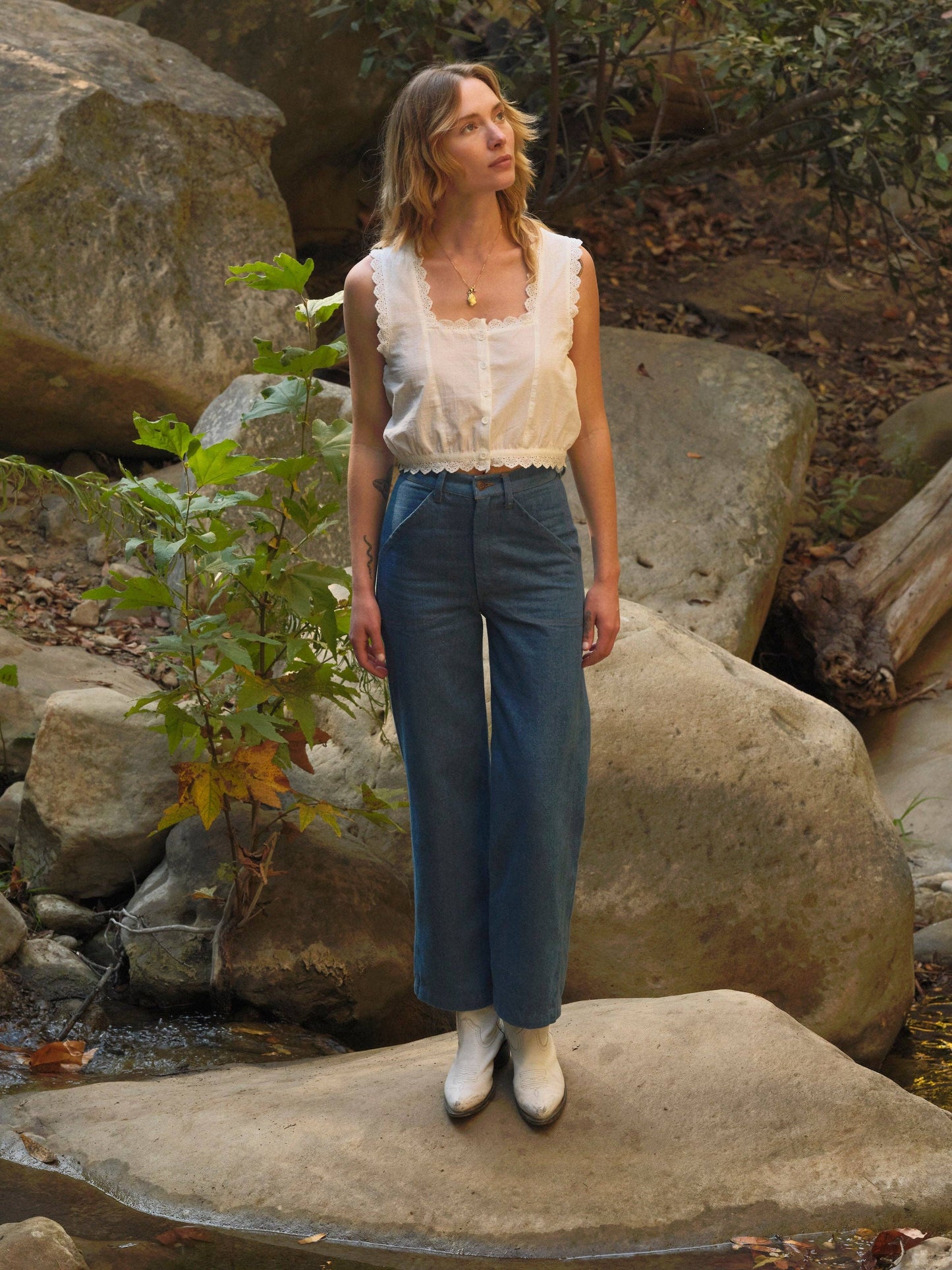 Woman in a light top, blue denim jeans, and white boots stands on rocky terrain outdoors.