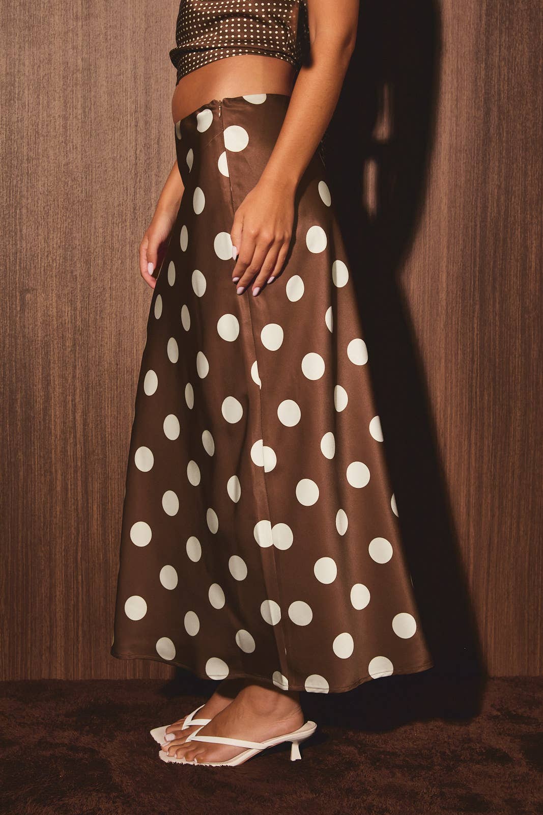 Brown polka dot skirt with studded crop top and white sandals, worn by a woman posing against a wooden wall.