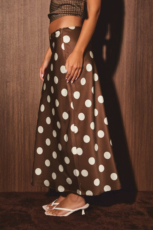 Brown polka dot skirt with studded crop top and white sandals, worn by a woman posing against a wooden wall.