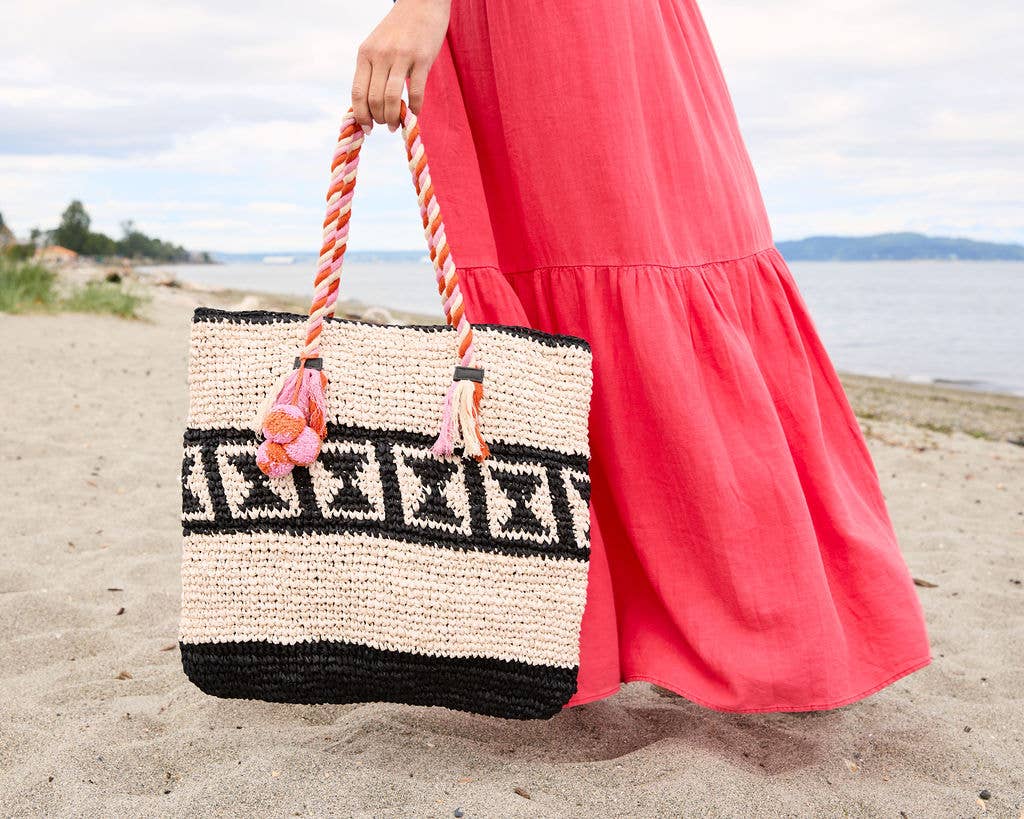 Woman in a pink dress carries a black-patterned raffia tote bag on the beach.