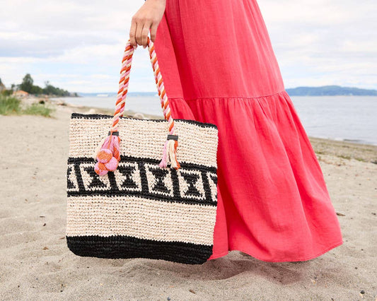 Woman in a pink dress carries a black-patterned raffia tote bag on the beach.