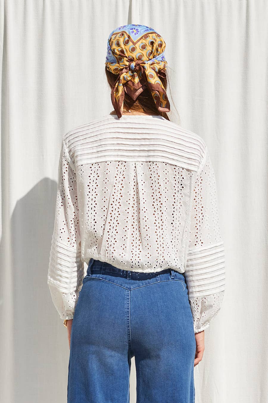 Woman in an off-white top, blue jeans, and a patterned headscarf stands facing away from the camera.