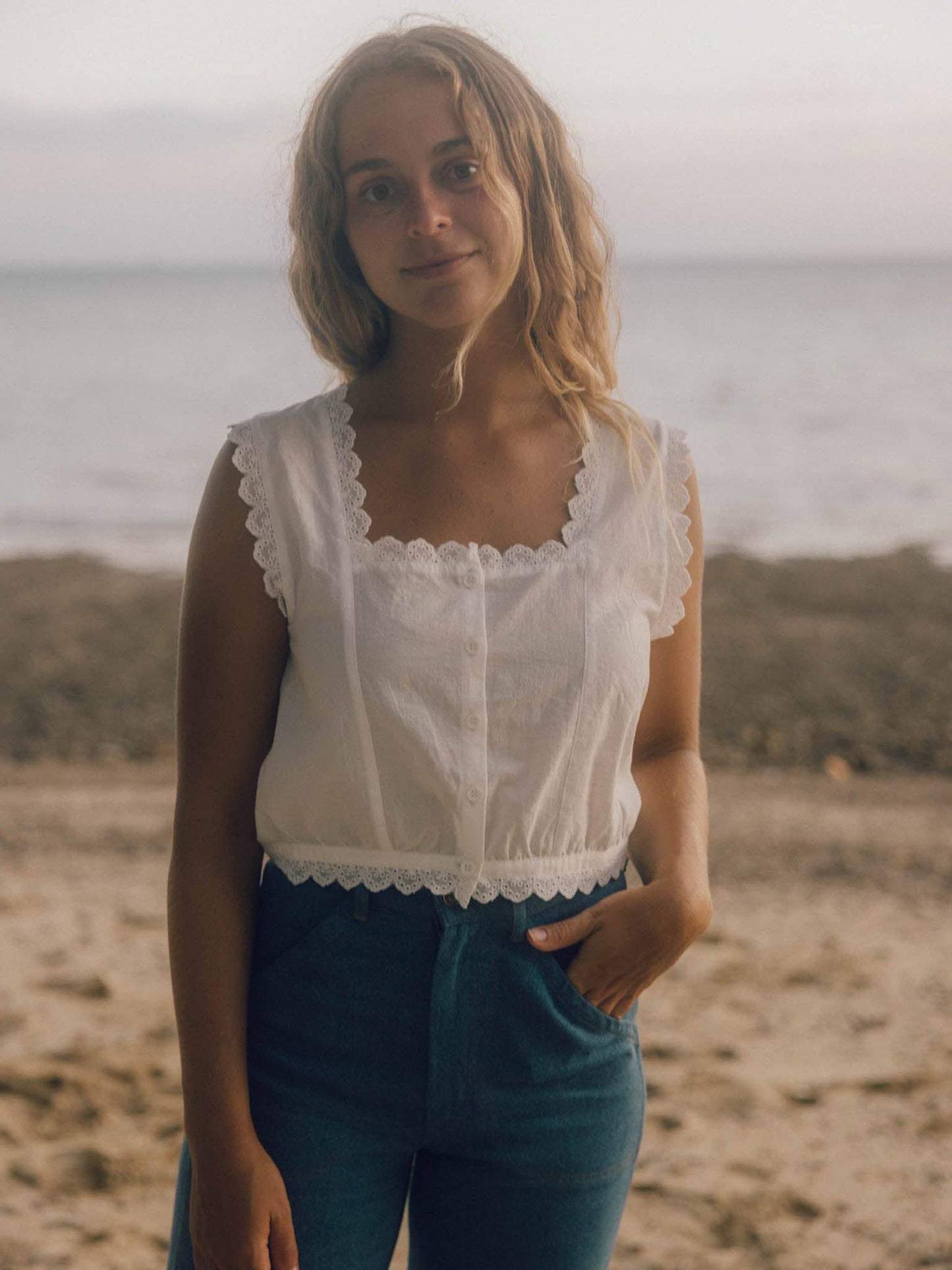 Woman in a relaxed linen top and blue denim jeans stands on a sandy beach, with the ocean visible behind her.