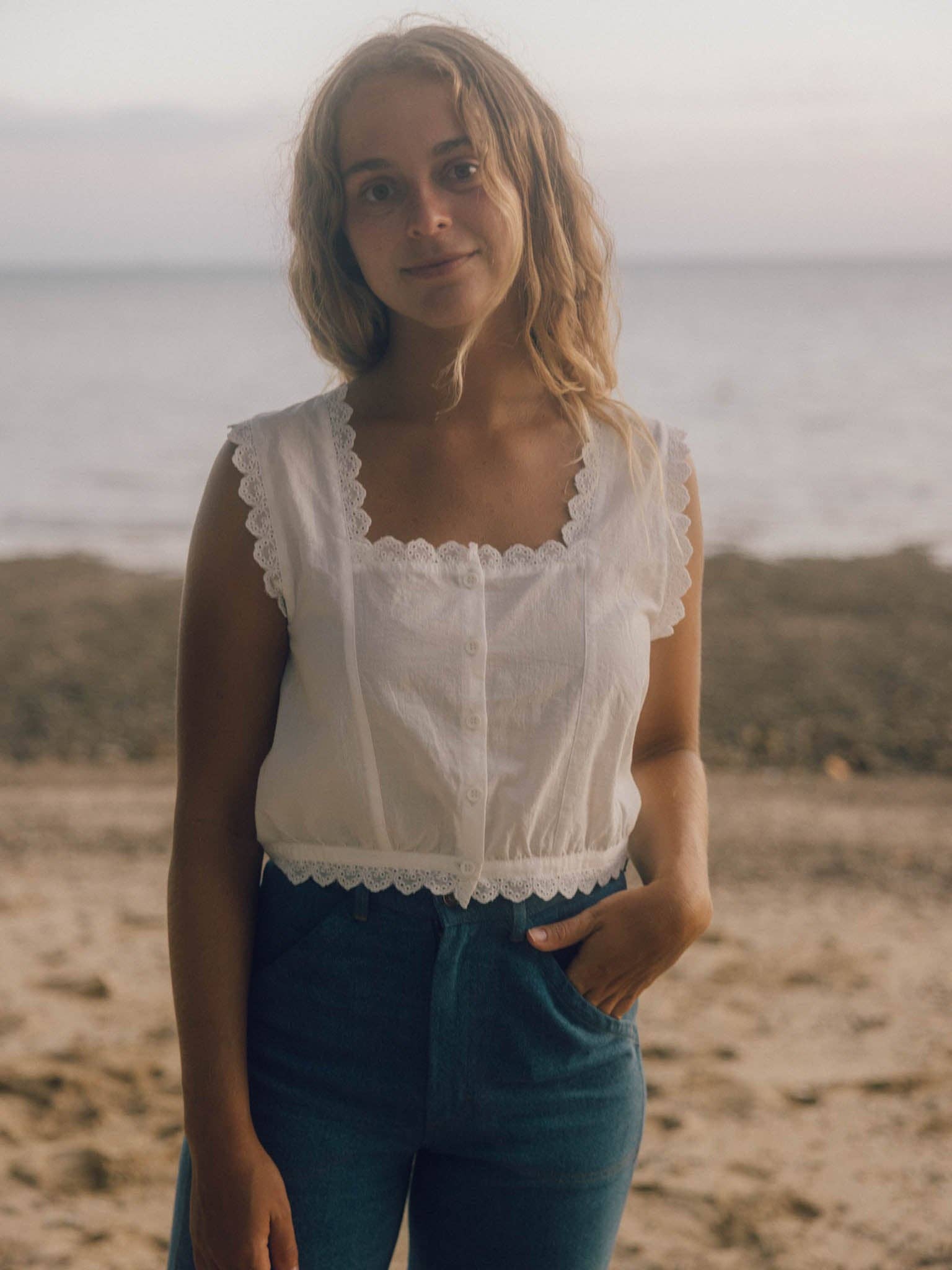 Woman in a relaxed linen top and blue denim jeans stands on a sandy beach, with the ocean visible behind her.