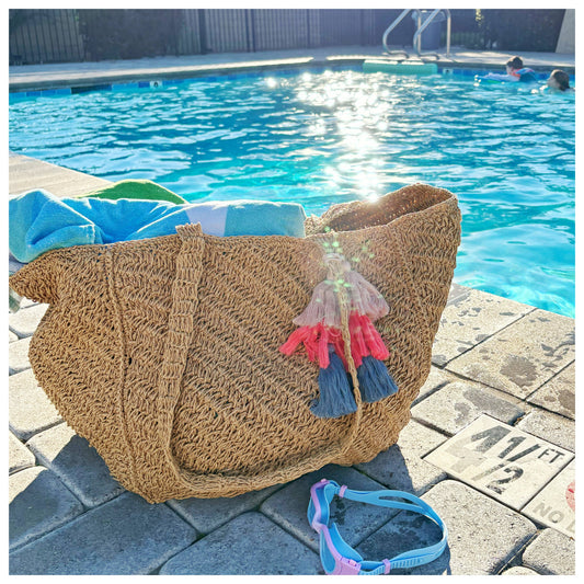 A woven canvas tote in neutral tones rests beside a sunlit pool, placed on the deck near people swimming.