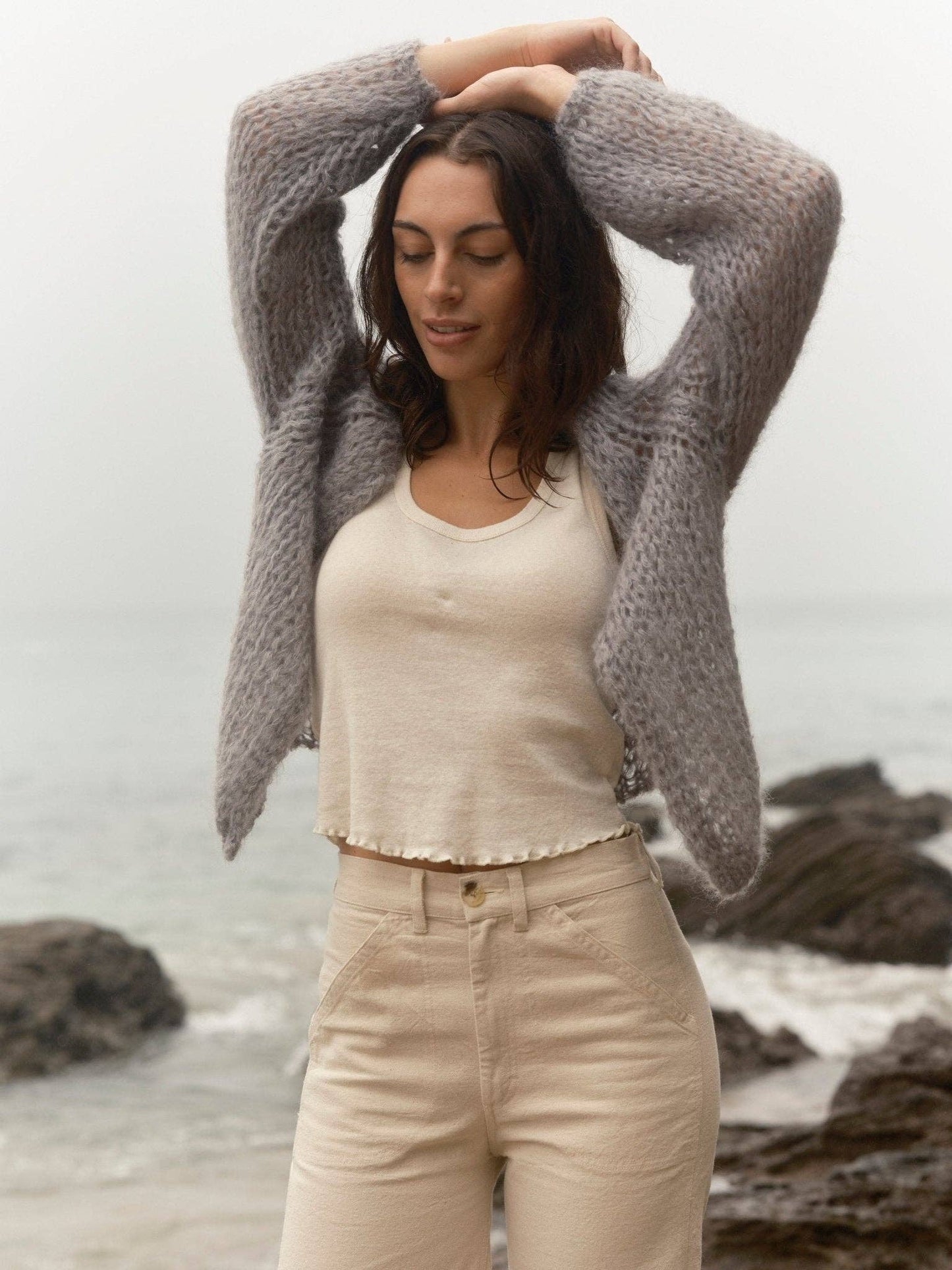 Woman in a cream knit cardigan stands on rocky shore, arms up, eyes closed—ideal for cool, casual outdoor wear.