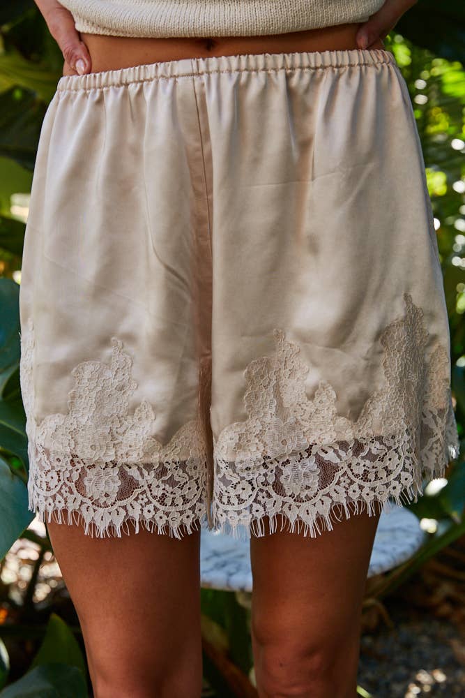 Cream lace-trim boho shorts, light fabric, styled for spring-summer, worn outdoors among green plants.