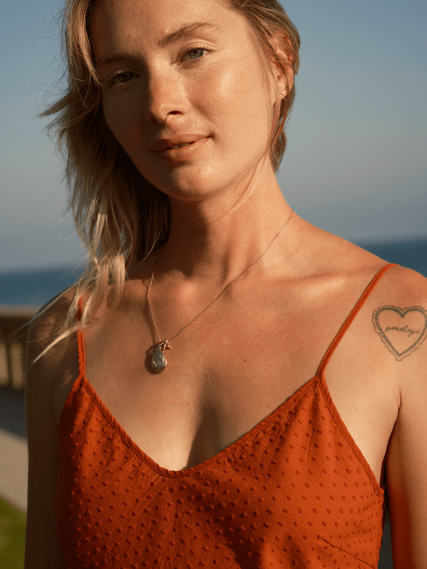 Woman outdoors in a light mollusk-colored dobby slip dress with necklace and heart tattoo on a sunny ocean day.