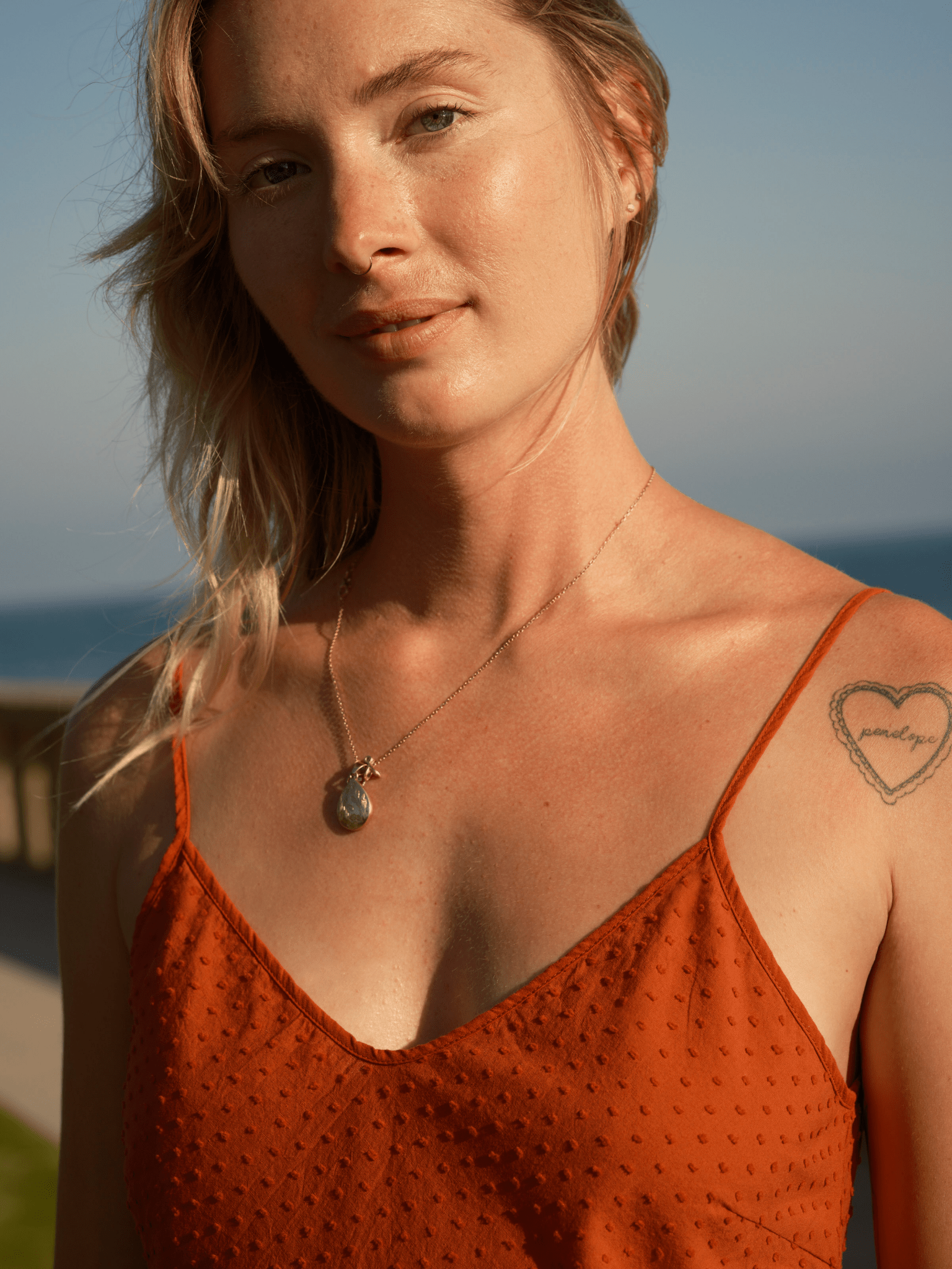 Woman outdoors in a light mollusk-colored dobby slip dress with necklace and heart tattoo on a sunny ocean day.