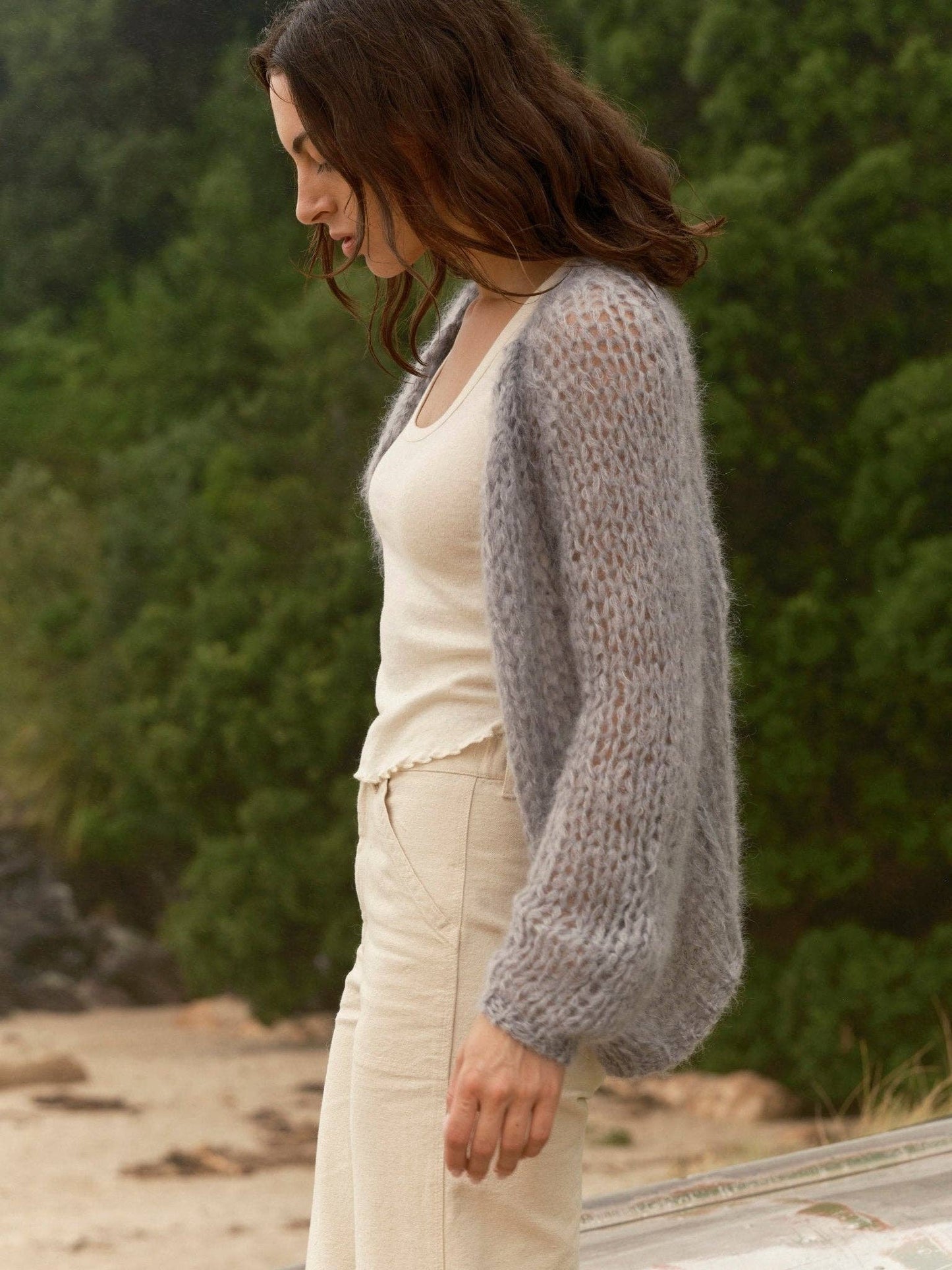 Woman wears cloud-patterned cardigan, beige pants, and tank top outdoors near trees and sand.