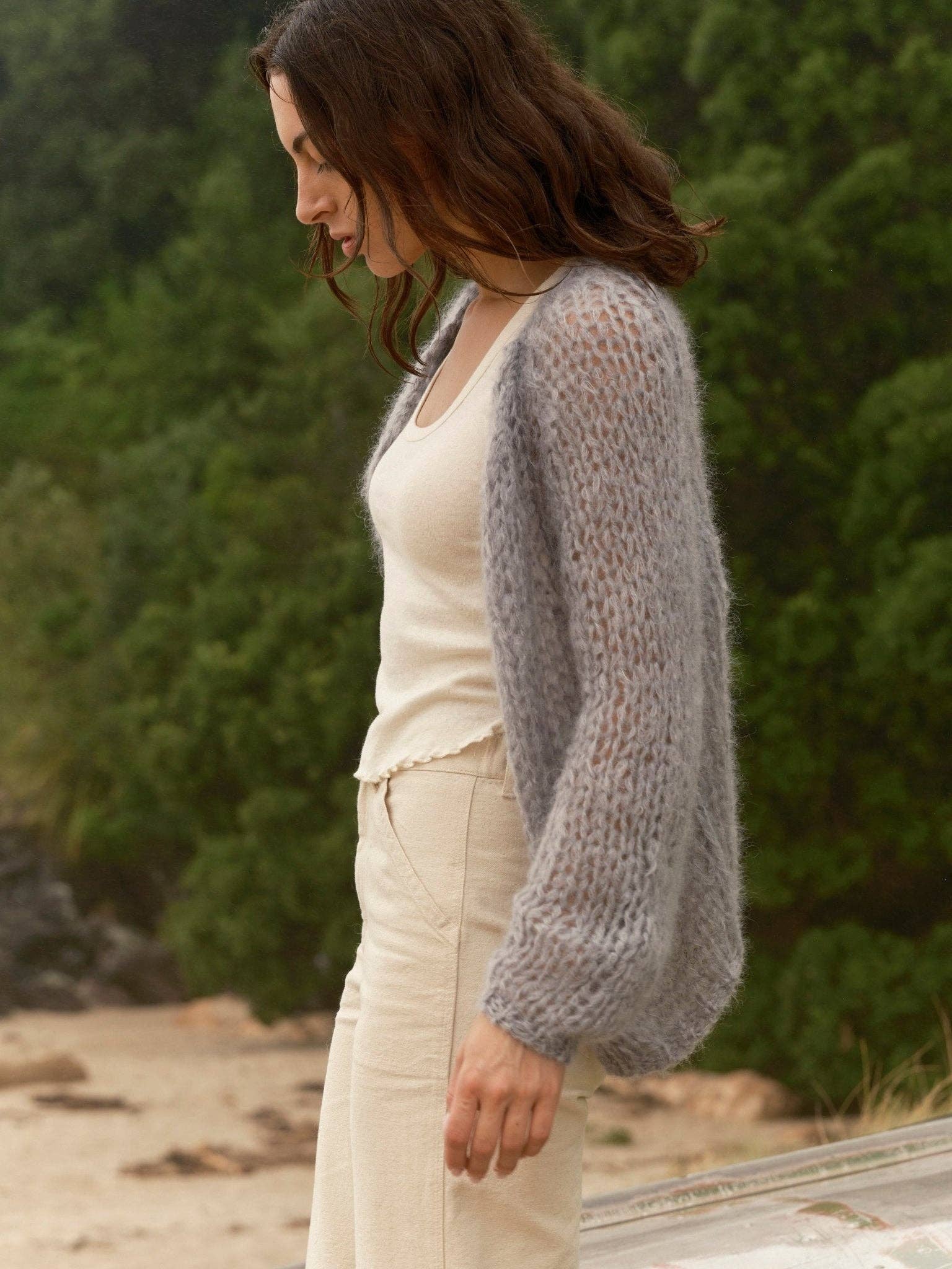 Woman wears cloud-patterned cardigan, beige pants, and tank top outdoors near trees and sand.