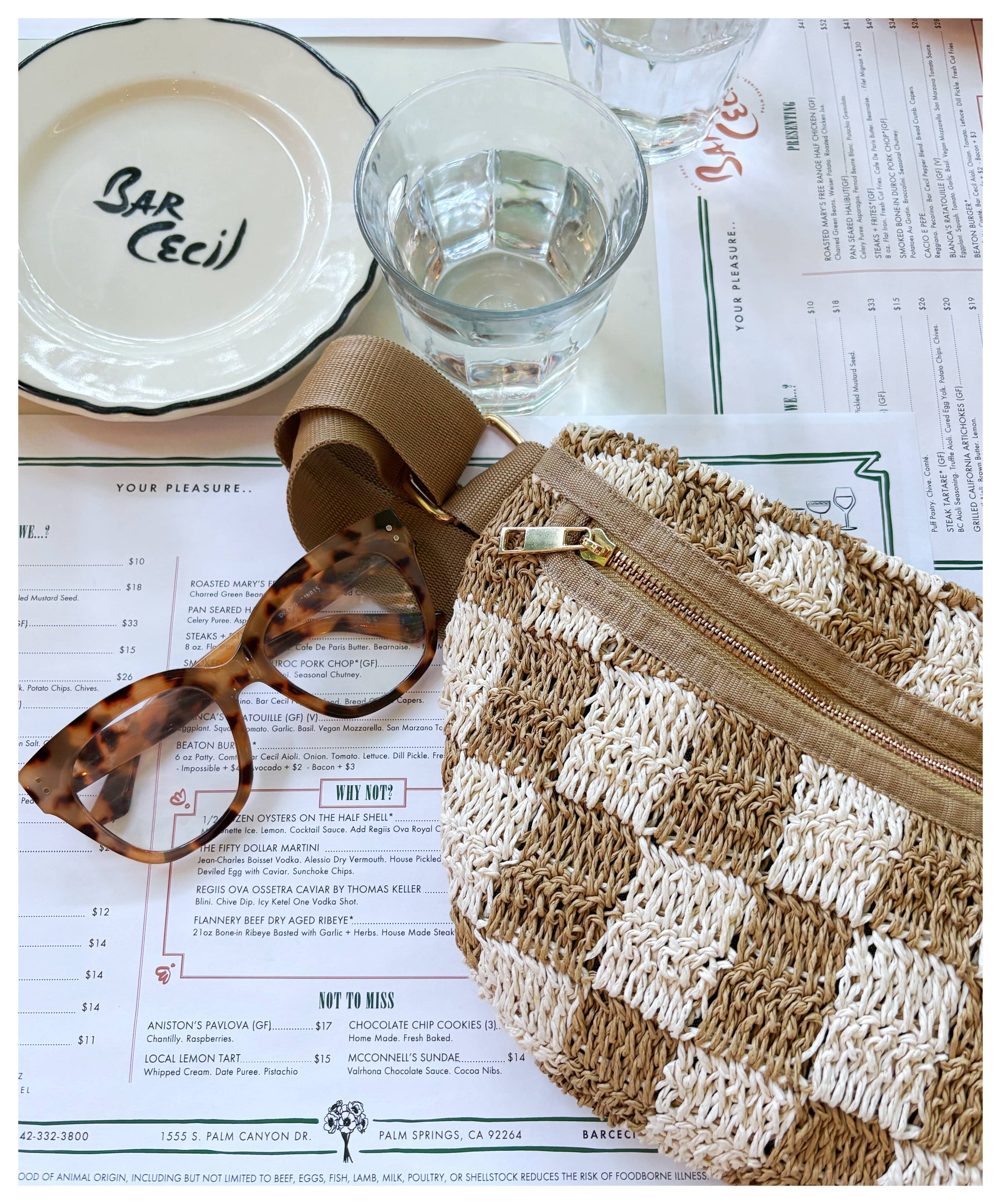 Checkered fabric sling bag, brown tortoiseshell glasses, and clear glassware on a plate beside a printed menu.