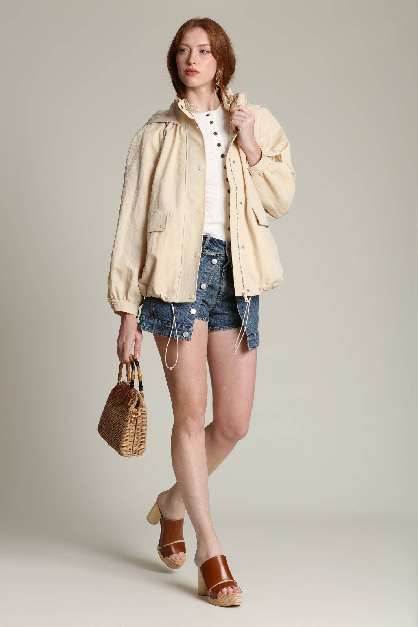 Woman in balloon-sleeve windbreaker, white shirt, denim shorts, brown sandals, and carrying a woven handbag.
