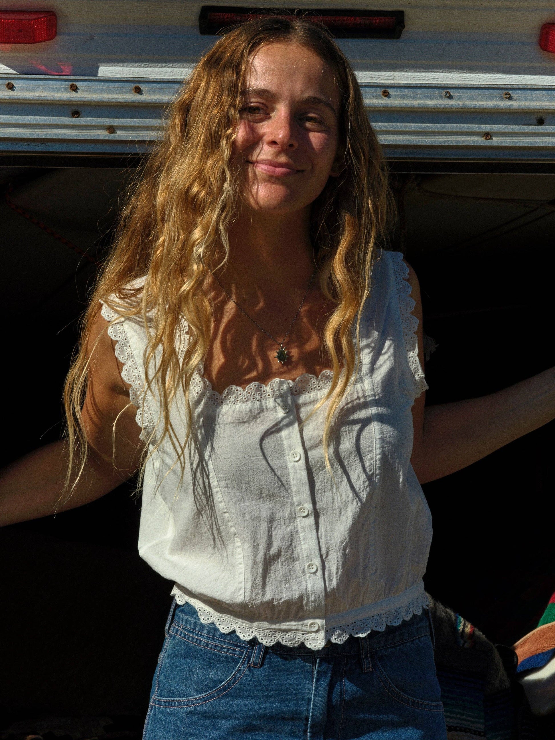 Woman outdoors in sunlight wears a light-colored sleeveless top and blue denim jeans, hair loose and wavy.