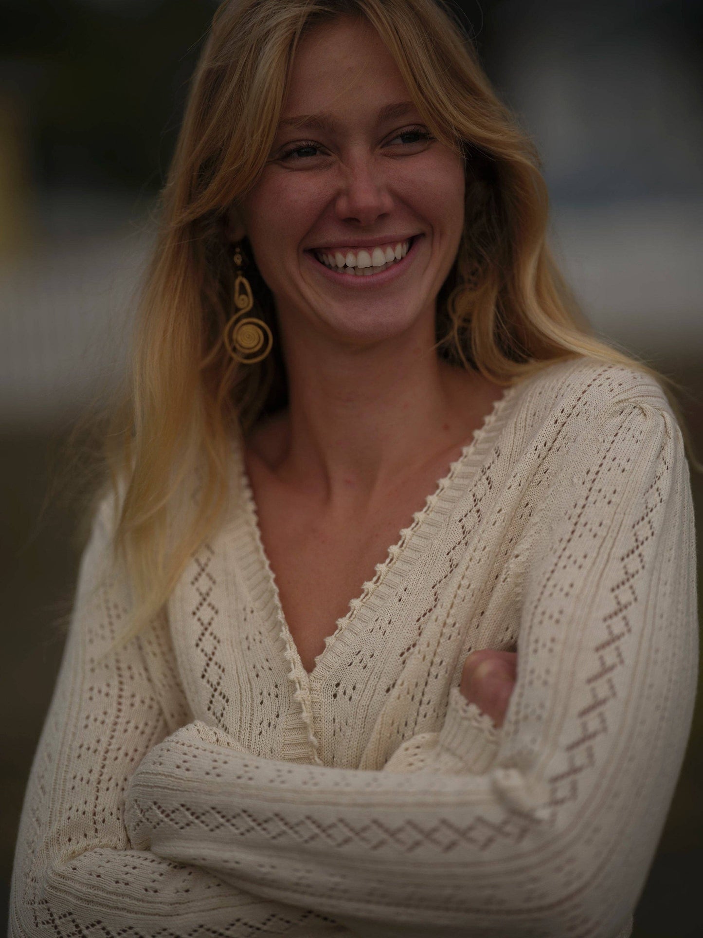 Woman outdoors wearing a cream knit cardigan and gold earrings, with long blonde hair.