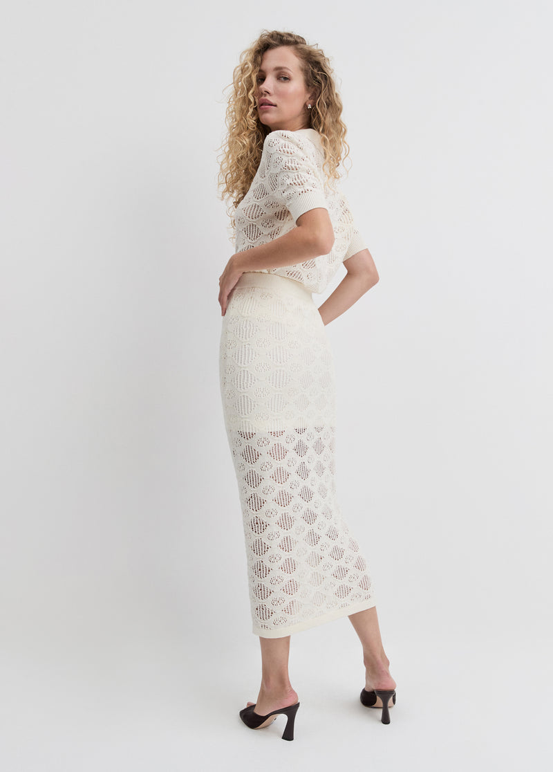 Curly-haired woman in an ivory pleated tennis skirt and white sweater, paired with black heels for a casual outing.