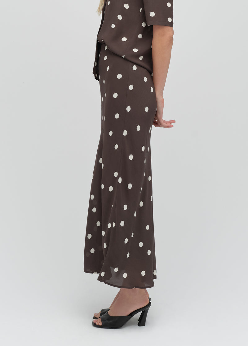 Woman in an espresso brown polka dot midi skirt and black heels stands sideways, showing flowy fabric and tailored fit.