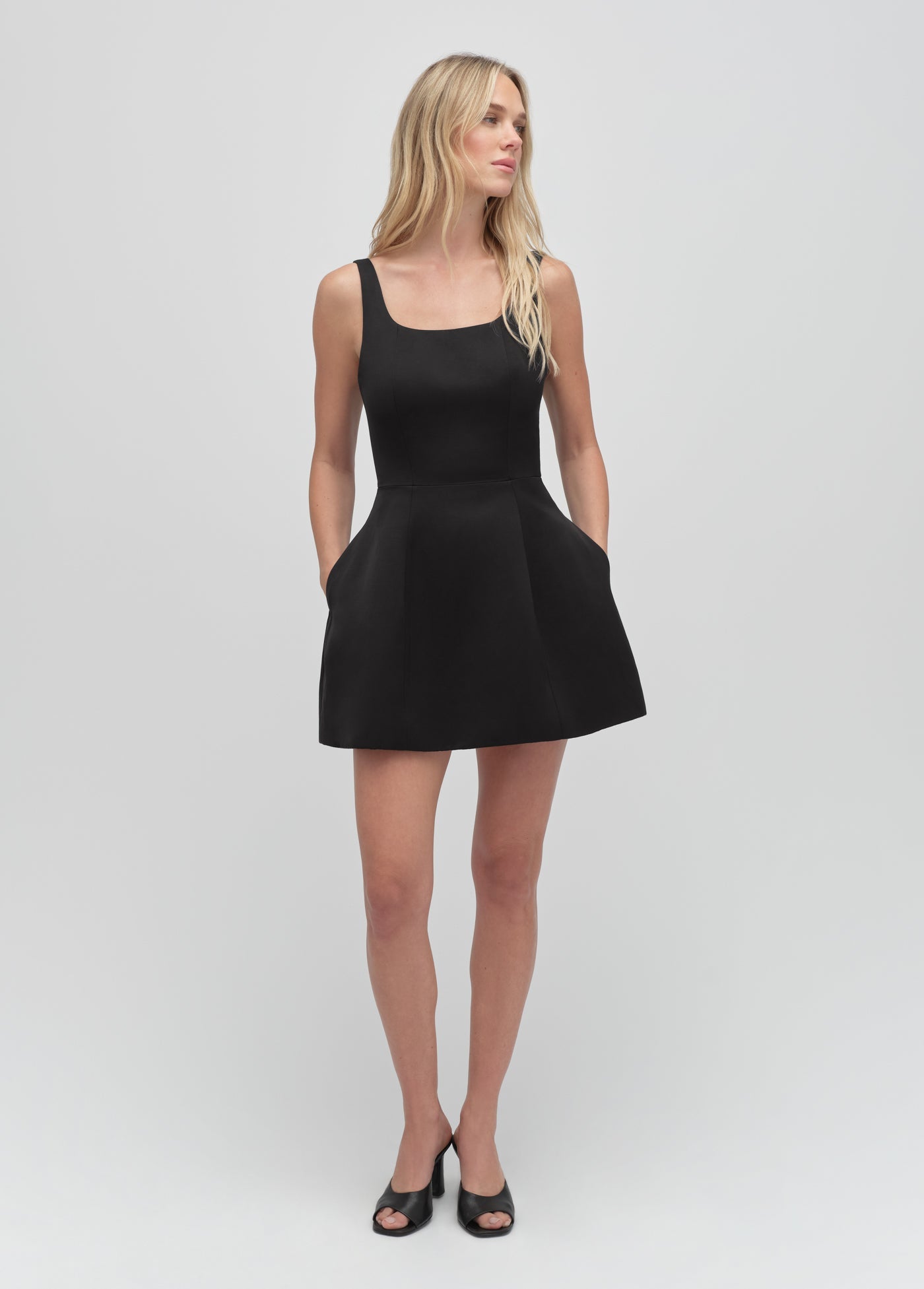 Woman in black midi dress and open-toe heels stands against a plain light background; suitable for formal or work occasions.