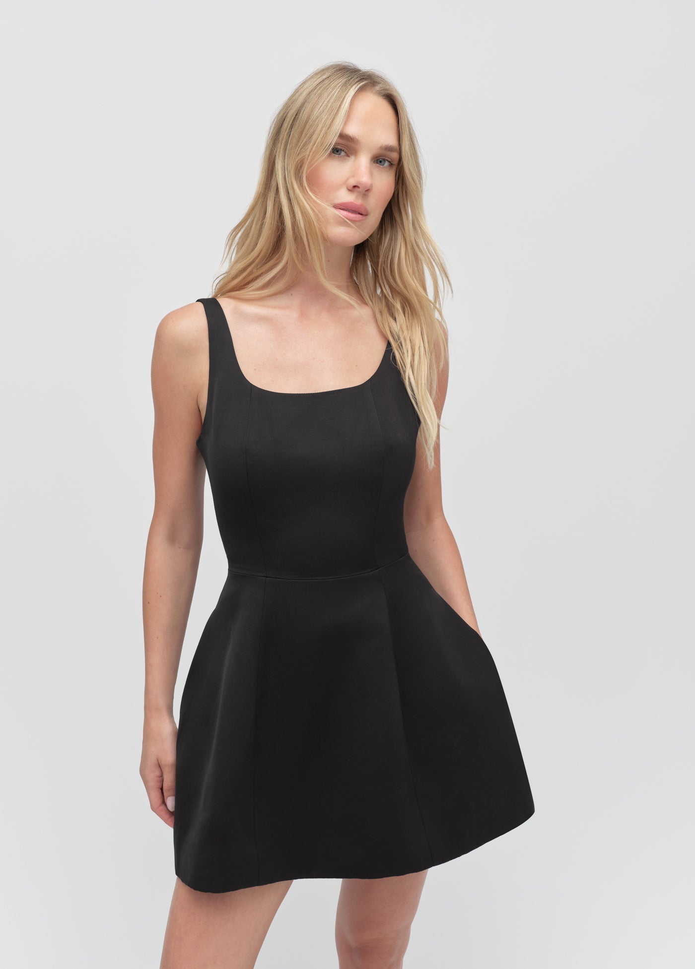 Sleeveless black mini dress with a scoop neck, shown on a model against a plain white background.