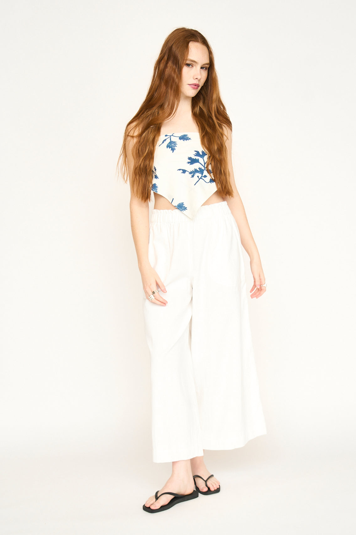 Woman with long red hair wears a floral bandana in Summer Fig, a patterned blouse, white pants, and black sandals.